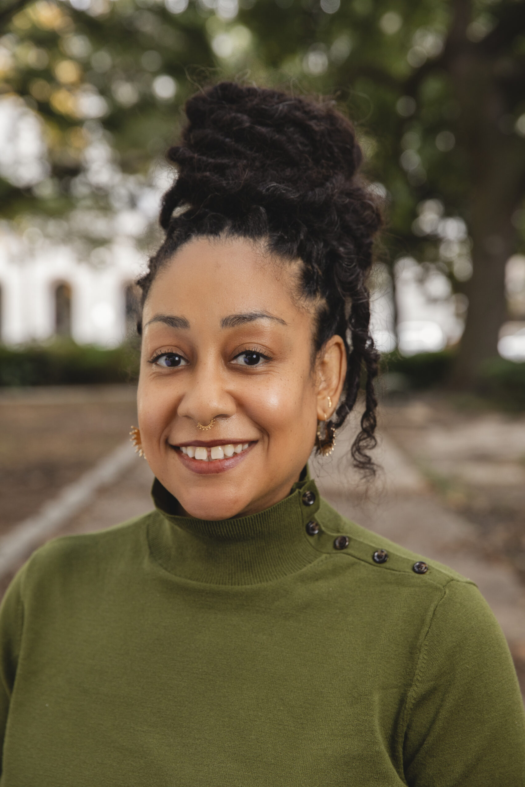 Headshot for Lauren Goudeaux. Lauren wears a green turtle-neck sweater with little black buttons along the neck. She has small gold earrings and a nose ring.