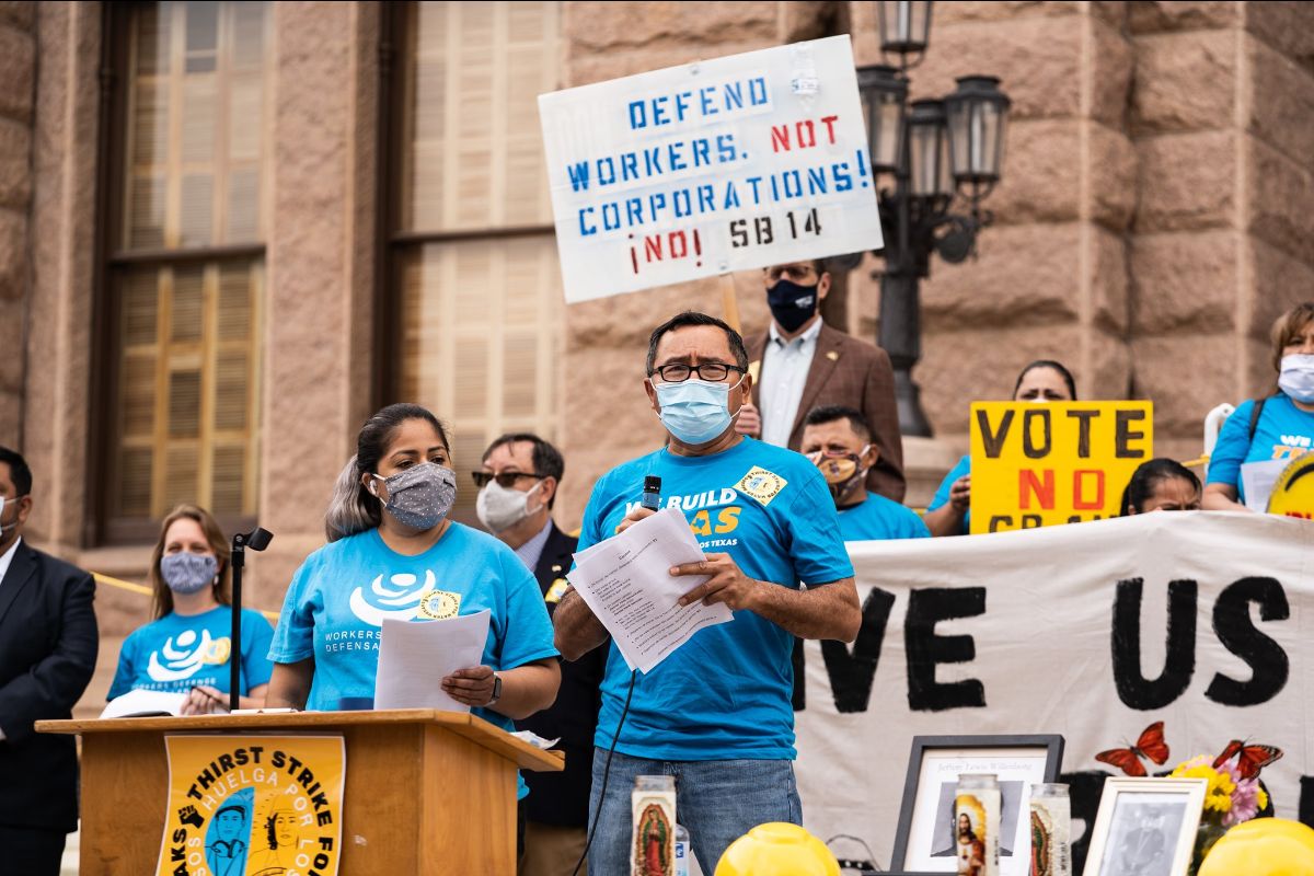 A photo of a group of a protestors wearing shirts from Workers Defense Project, standing at a podium and carrying a sign that says, A photo of a group of a protestors wearing shirts from Workers Defense Project, standing at a podium and carrying a sign that says,