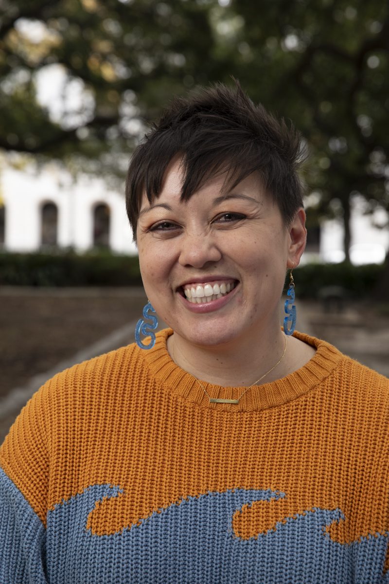 Headshot for Courtney Banayad. Courtney is wearing a bright orange and light blue sweater with blue earrings that have a squiggly shape.