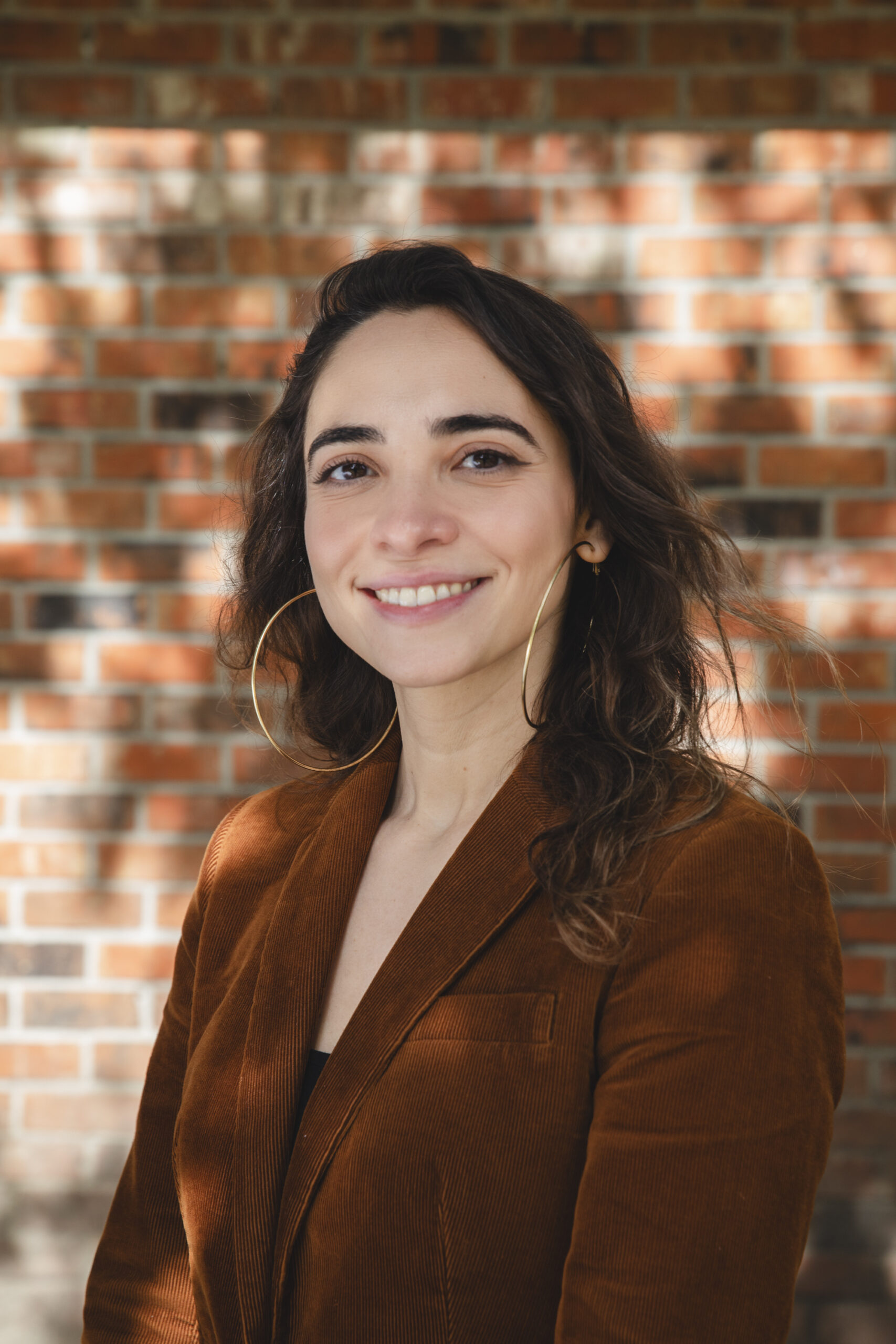 Headshot for Renata Peralta. Renata wears gold hoops and a copper blazer. She stands in front of a brick wall.