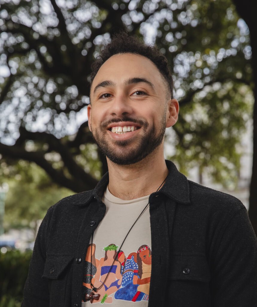 Headshot of Stephan Cheney. Stephan stand in front a large tree wearing a dark shirt jacket over a light grey graphic t-shirt with a dragonfly carved wood necklace.