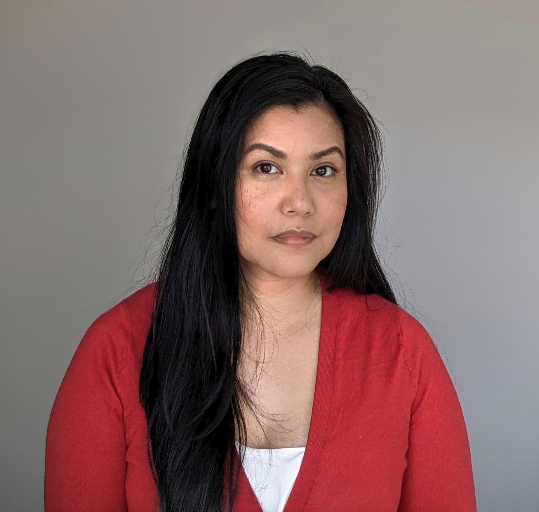 Headshot for Isabel Bombaes. Isabel sits in front of a grey background and wears a bright red sweater with a white shirt underneath.