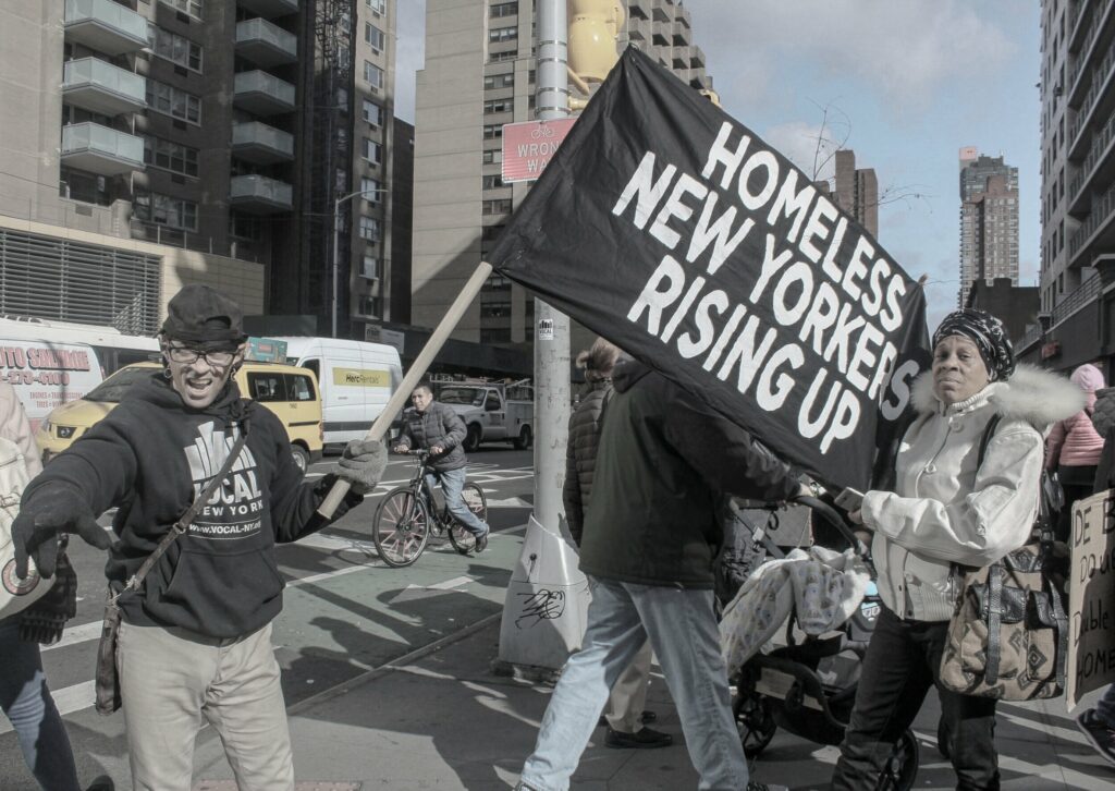 Photo of people outside at a protest with an organizer on the left holding a banner with black ground and white text that reads, 'Homeless New Yorkers Rising Up'