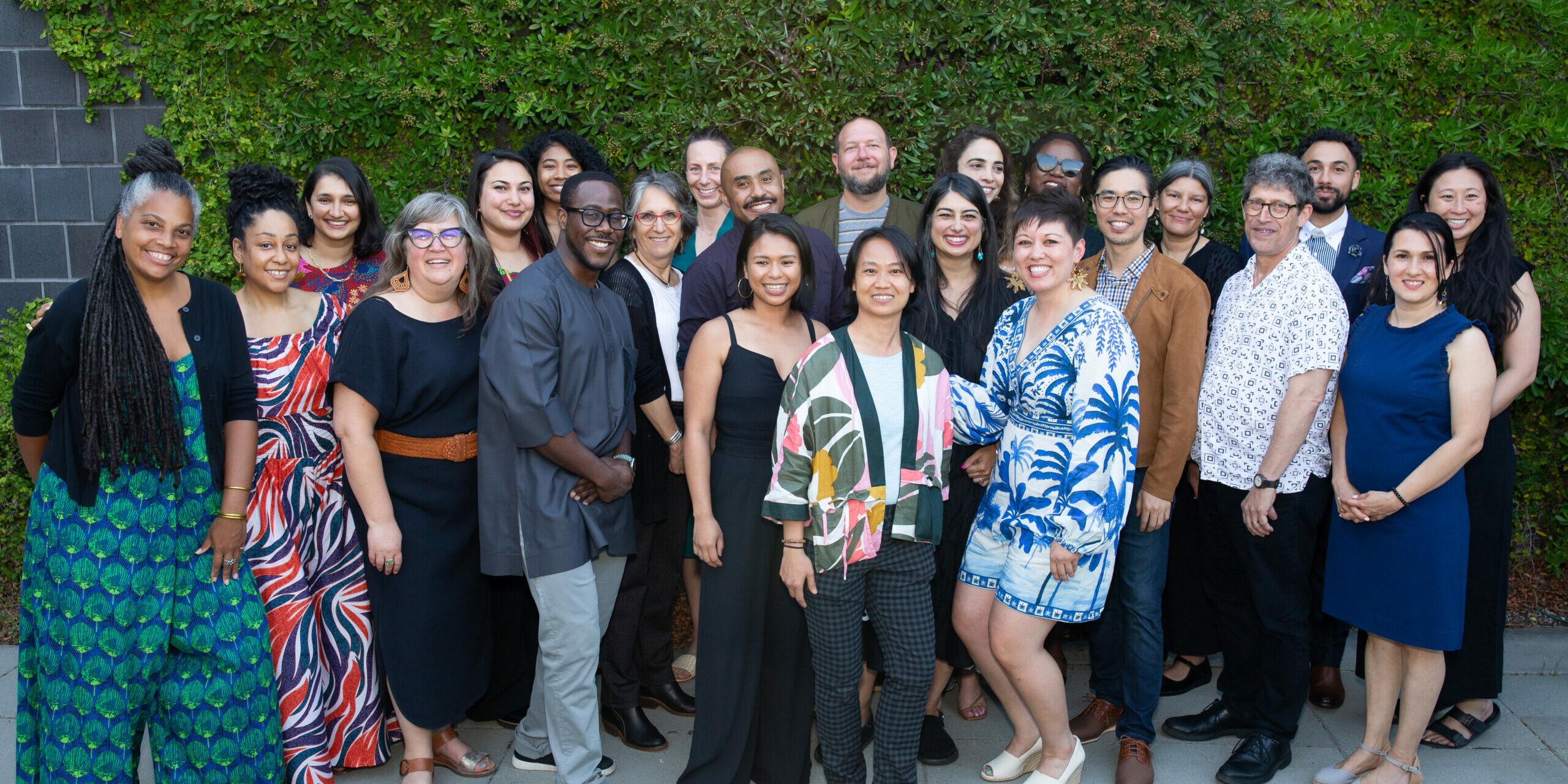 NFG staff and board gather for a group photo in front of a brick wall covered in lush greenery at the Cameron Art Museum.